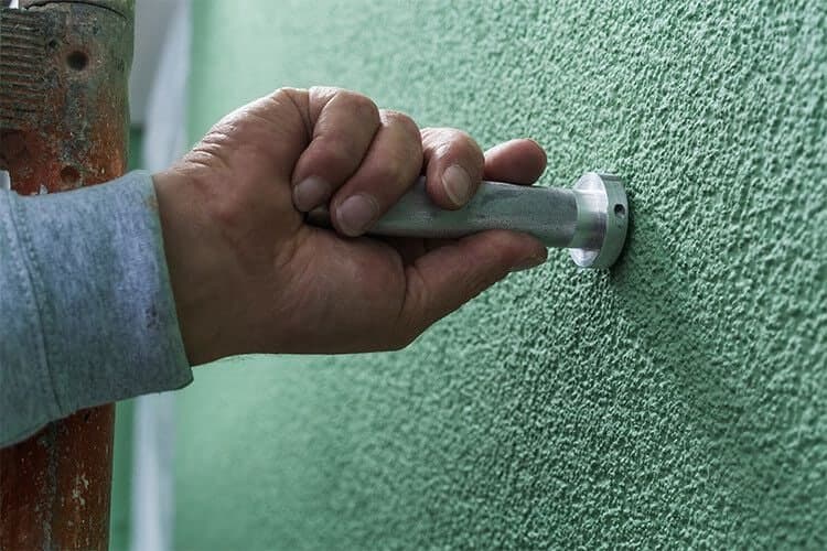Eine Hand führt den Edelstahl-Handfräser in das Gerüstankerloch einer grün verputzten Fassade.
